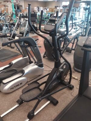 Gym with various cardio equipment, including a black elliptical machine and a silver elliptical in the foreground.